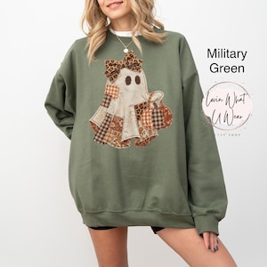 Op de afbeelding: Een militair groen sweatshirt met ronde hals en een afbeelding van een geest met een strik met luipaardprint die een pompoen vasthoudt. De geest heeft patchworkpatronen in bruin, oranje en wit. Tekst rechts: Military Green en Lovin What U Wear.