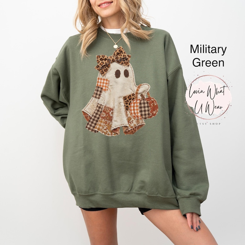 Op de afbeelding: Een militair groen sweatshirt met ronde hals en een afbeelding van een geest met een strik met luipaardprint die een pompoen vasthoudt. De geest heeft patchworkpatronen in bruin, oranje en wit. Tekst rechts: Military Green en Lovin What U Wear.