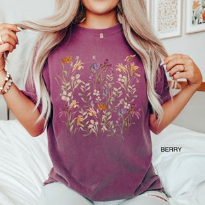 May include: A purple t-shirt with a floral print featuring a variety of flowers in shades of yellow, pink, and purple. The flowers are arranged in a cluster on the front of the shirt.