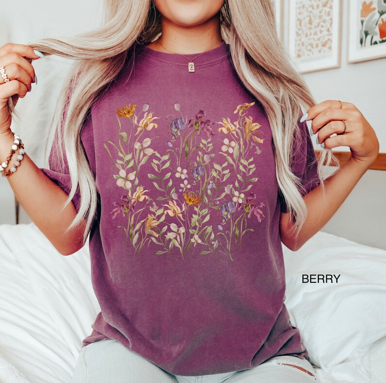 May include: A purple t-shirt with a floral print featuring a variety of flowers in shades of yellow, pink, and purple. The flowers are arranged in a cluster on the front of the shirt.