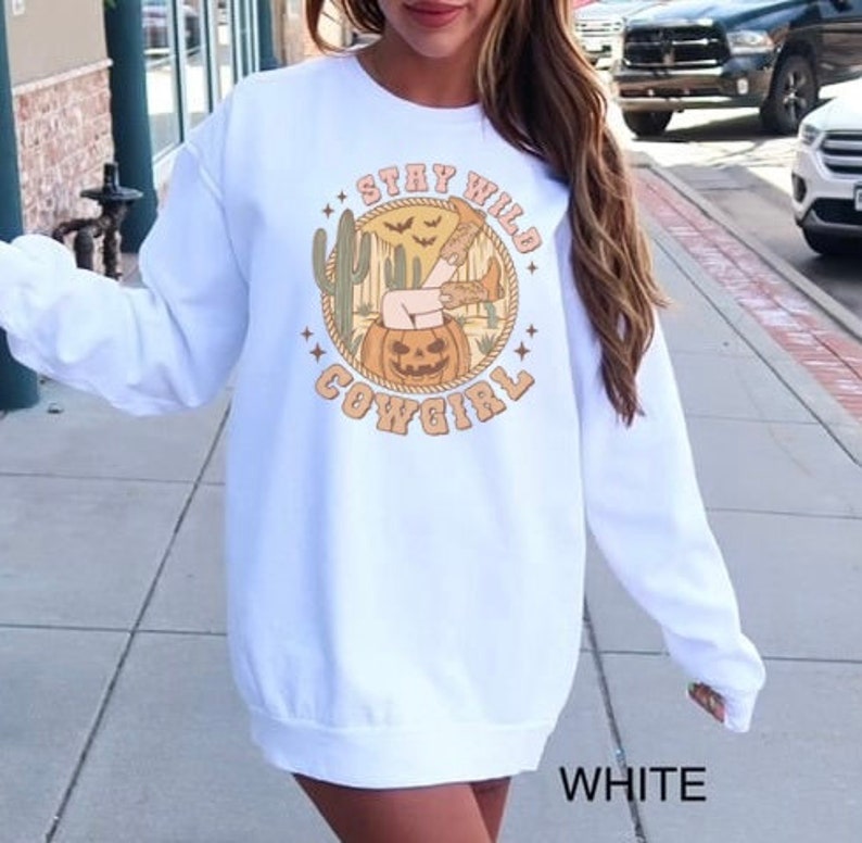 May include: White long-sleeved sweatshirt with a graphic print of a woman's legs sticking out of a jack-o-lantern. The graphic also includes the text "Stay Wild Cowgirl" in a vintage style font.