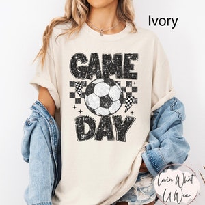 May include: Ivory-colored t-shirt with a distressed "GAME DAY" graphic featuring a soccer ball, checkered flags, and stars. The shirt is a casual, unisex style, perfect for sports fans.