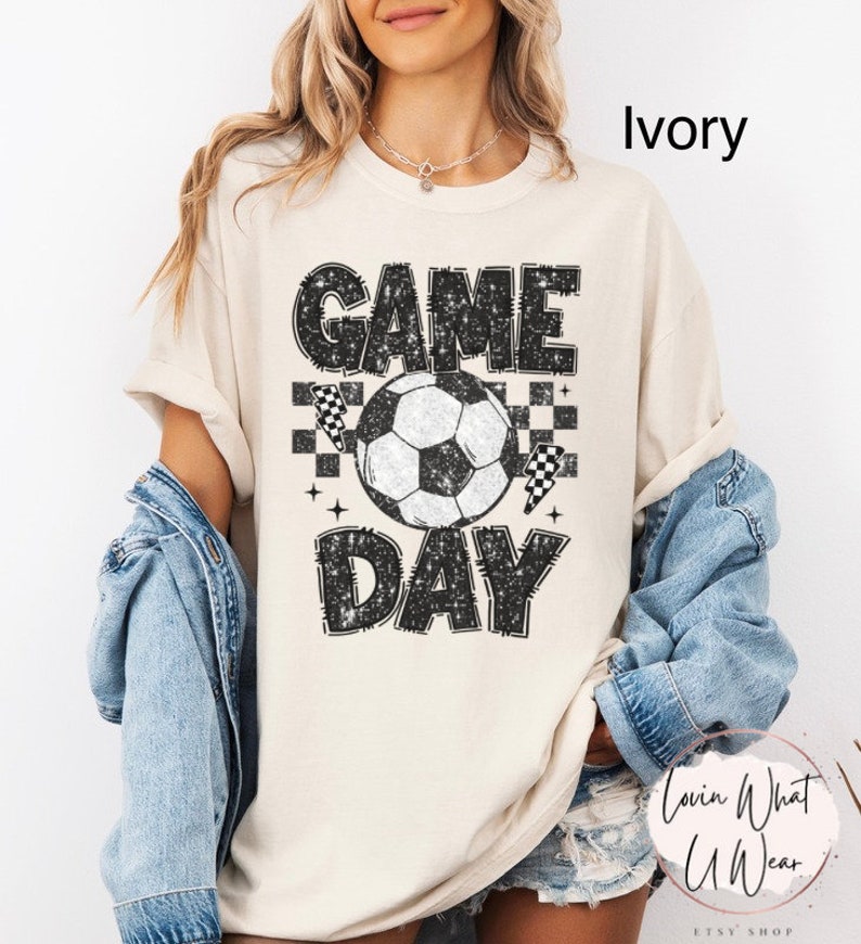 May include: Ivory-colored t-shirt with a distressed "GAME DAY" graphic featuring a soccer ball, checkered flags, and stars. The shirt is a casual, unisex style, perfect for sports fans.