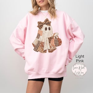 Op de afbeelding: Een lichtroze sweater met ronde hals en een centrale afbeelding van een geest met een strik met luipaardprint. De geest is samengesteld uit verschillende bruine, oranje en witte stoffen met patronen en houdt twee kleine pompoenen vast.