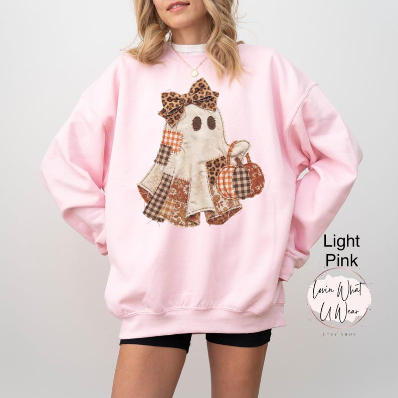 Op de afbeelding: Een lichtroze sweater met ronde hals en een centrale afbeelding van een geest met een strik met luipaardprint. De geest is samengesteld uit verschillende bruine, oranje en witte stoffen met patronen en houdt twee kleine pompoenen vast.