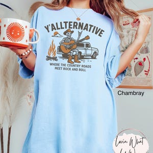 May include: Light blue t-shirt with the text "Y'ALLTERNATIVE" and a graphic of a skeleton playing a guitar next to a truck and campfire. The phrase "WHERE THE COUNTRY ROADS MEET ROCK AND ROLL" is below the graphic.