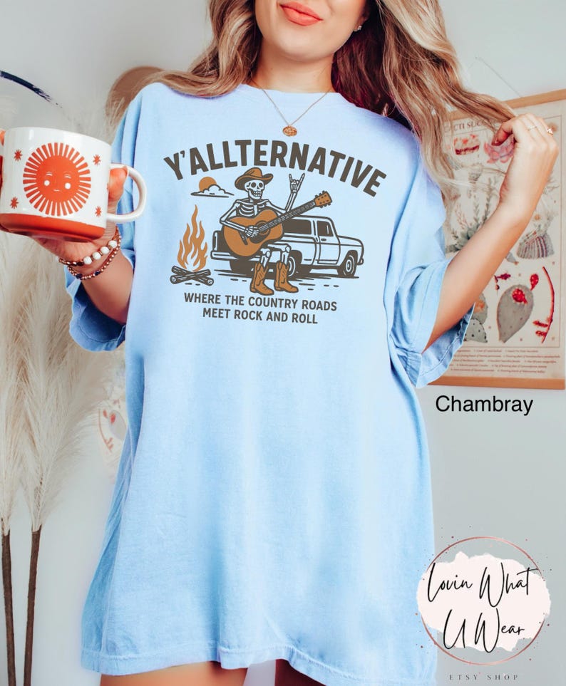May include: Light blue t-shirt with the text "Y'ALLTERNATIVE" and a graphic of a skeleton playing a guitar next to a truck and campfire. The phrase "WHERE THE COUNTRY ROADS MEET ROCK AND ROLL" is below the graphic.
