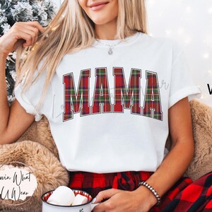 May include: White t-shirt with the word "MAMA" in a red, green, and white plaid design. The shirt is part of a holiday-themed collection. The model is wearing a bracelet and holding a mug.
