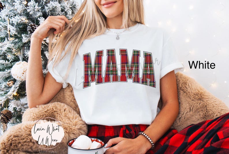 May include: White t-shirt with the word "MAMA" in a red, green, and white plaid design. The shirt is part of a holiday-themed collection. The model is wearing a bracelet and holding a mug.