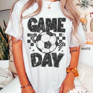 May include: White t-shirt with a distressed black graphic that reads "GAME DAY" above and below a soccer ball. The design includes checkered flags and stars. The shirt is a casual style.