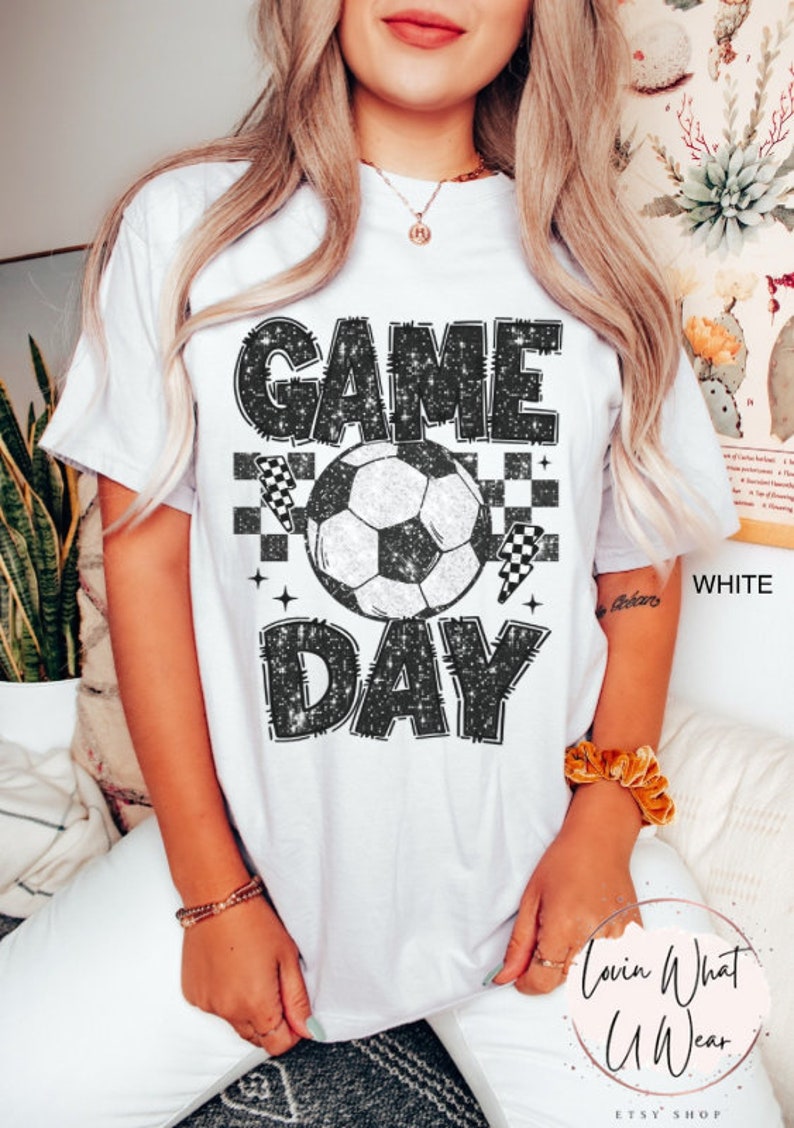 May include: White t-shirt with a distressed black graphic that reads "GAME DAY" above and below a soccer ball. The design includes checkered flags and stars. The shirt is a casual style.