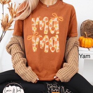 May include: A burnt orange t-shirt with the word "Mama" in a pumpkin-themed design. The letters are filled with pumpkin illustrations and accented with orange bows. The shirt is part of a fall-themed collection.