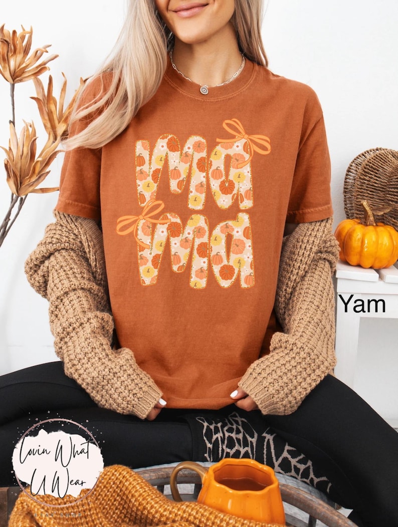 May include: A burnt orange t-shirt with the word "Mama" in a pumpkin-themed design. The letters are filled with pumpkin illustrations and accented with orange bows. The shirt is part of a fall-themed collection.