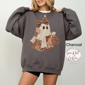 Op de afbeelding: Een antracietgrijs sweatshirt met lange mouwen en een centrale afbeelding van een geest met een strik met luipaardprint die een pompoen vasthoudt. De geest is versierd met diverse bruine en oranje ruitjes- en bloemenstoffen. Tekst Charcoal.