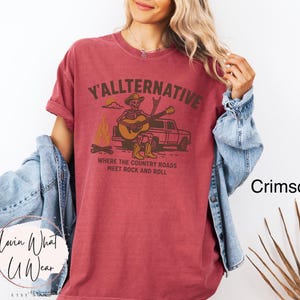 May include: Crimson-colored t-shirt with the text "Y'allternative" and a graphic of a skeleton playing guitar by a campfire and truck. The shirt's design includes the phrase "Where the country roads meet rock and roll."