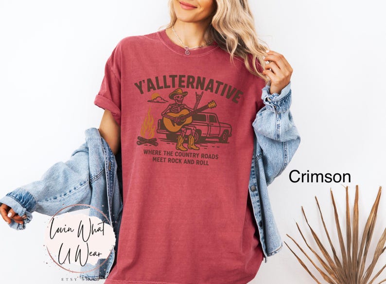 May include: Crimson-colored t-shirt with the text "Y'allternative" and a graphic of a skeleton playing guitar by a campfire and truck. The shirt's design includes the phrase "Where the country roads meet rock and roll."