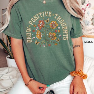 Op de afbeelding: Groen T-shirt met een vintage bloemenontwerp en de tekst "Grow Positive Thoughts".