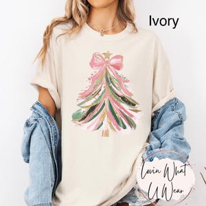 Op de afbeelding: Ivoor kleurig T-shirt met een kerstboom ontwerp. De boom is versierd met roze, groene en gouden penseelstreken, bekroond met een roze strik en een gouden ster. De tekst "Ivory" is op het shirt gedrukt.