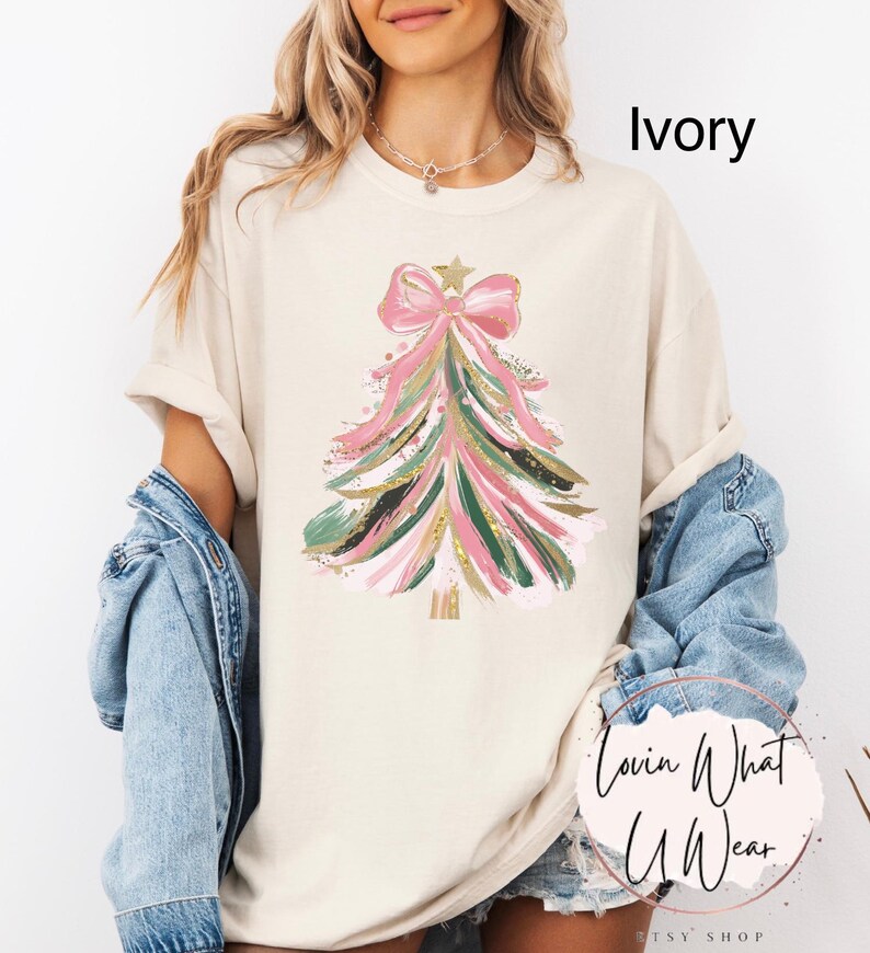 Op de afbeelding: Ivoor kleurig T-shirt met een kerstboom ontwerp. De boom is versierd met roze, groene en gouden penseelstreken, bekroond met een roze strik en een gouden ster. De tekst "Ivory" is op het shirt gedrukt.