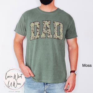 May include: Moss green t-shirt with the word "DAD" in a camouflage pattern across the chest. The shirt has short sleeves and a crew neck. The text "Lovin What U Wear" is at the bottom left.