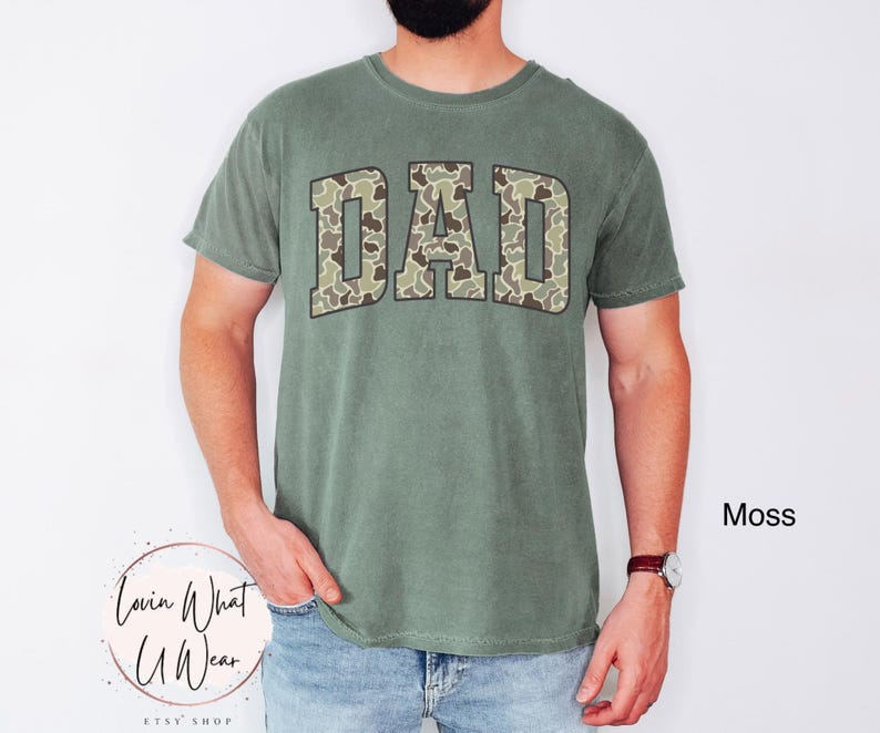 May include: Moss green t-shirt with the word "DAD" in a camouflage pattern across the chest. The shirt has short sleeves and a crew neck. The text "Lovin What U Wear" is at the bottom left.