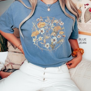 May include: A woman wearing a blue jean t-shirt with a floral print and white jeans. The t-shirt has a gold circle around the floral print. The text "BLUE JEAN" is in the bottom right corner of the image.