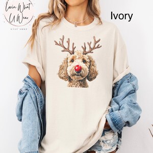 K&ouml;nnte beinhalten: Ein T-Shirt in Elfenbein mit einem braunen Hund mit Rentiergeweih und einer roten Nase. Der Text "Lovin What U Wear" befindet sich in einem Kreis &uuml;ber dem Hund.