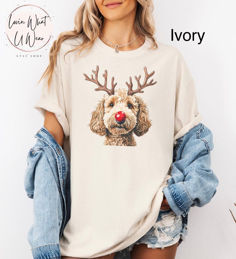 K&ouml;nnte beinhalten: Ein T-Shirt in Elfenbein mit einem braunen Hund mit Rentiergeweih und einer roten Nase. Der Text "Lovin What U Wear" befindet sich in einem Kreis &uuml;ber dem Hund.