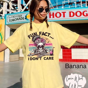 May include: Yellow t-shirt with a skeleton wearing sunglasses and a flower crown. The text on the shirt reads "Fun Fact... I Don't Care".