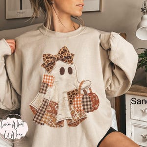 Op de afbeelding: Een zandkleurige sweater met ronde hals en een centrale afbeelding van een geest versierd met een strik met luipaardprint. De geest is samengesteld uit diverse stoffen patches in oranje, bruin en wit met ruitjes, bloemen en luipaardpatronen.