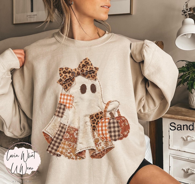 Op de afbeelding: Een zandkleurige sweater met ronde hals en een centrale afbeelding van een geest versierd met een strik met luipaardprint. De geest is samengesteld uit diverse stoffen patches in oranje, bruin en wit met ruitjes, bloemen en luipaardpatronen.