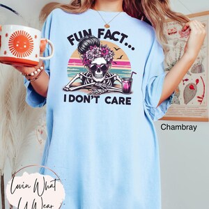 May include: Light blue t-shirt with a graphic of a skeleton wearing sunglasses and a flower crown. The graphic is set against a sunset background with the text "Fun Fact... I Don't Care".