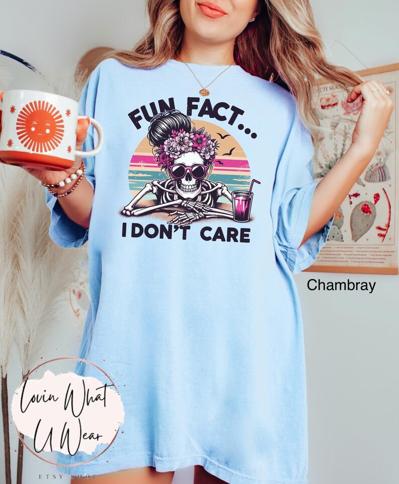May include: Light blue t-shirt with a graphic of a skeleton wearing sunglasses and a flower crown. The graphic is set against a sunset background with the text "Fun Fact... I Don't Care".