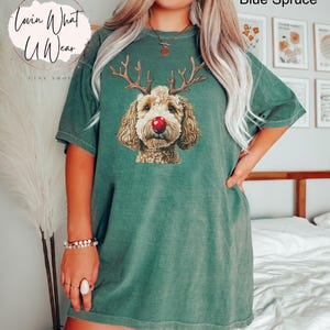 K&ouml;nnte beinhalten: Ein gr&uuml;nes T-Shirt mit einem braunen und wei&szlig;en Hund, der eine rote Nase und Rentiergeweih tr&auml;gt. Der Text "Lovin What U Wear" befindet sich in der oberen linken Ecke des Bildes.