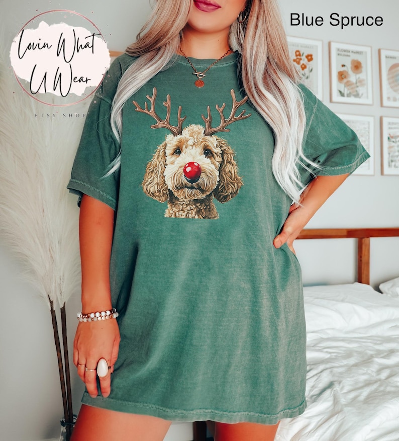 K&ouml;nnte beinhalten: Ein gr&uuml;nes T-Shirt mit einem braunen und wei&szlig;en Hund, der eine rote Nase und Rentiergeweih tr&auml;gt. Der Text "Lovin What U Wear" befindet sich in der oberen linken Ecke des Bildes.