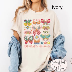 May include: A cream colored t-shirt with a colorful butterfly design and the text "Be Kind to all kinds". The shirt is being modeled by a person wearing a denim jacket.