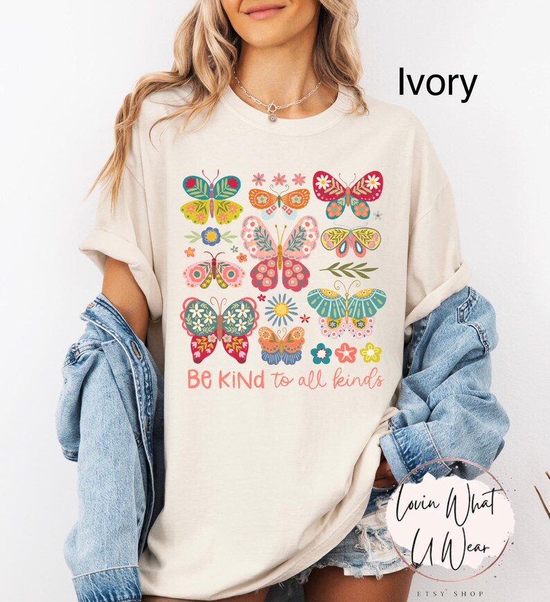 May include: A cream colored t-shirt with a colorful butterfly design and the text "Be Kind to all kinds". The shirt is being modeled by a person wearing a denim jacket.