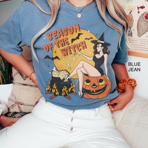 Puede incluir: Una camiseta azul con una ilustraci&oacute;n vintage de una bruja sentada sobre una calabaza. El texto "Season of the Witch" est&aacute; impreso en la camiseta.