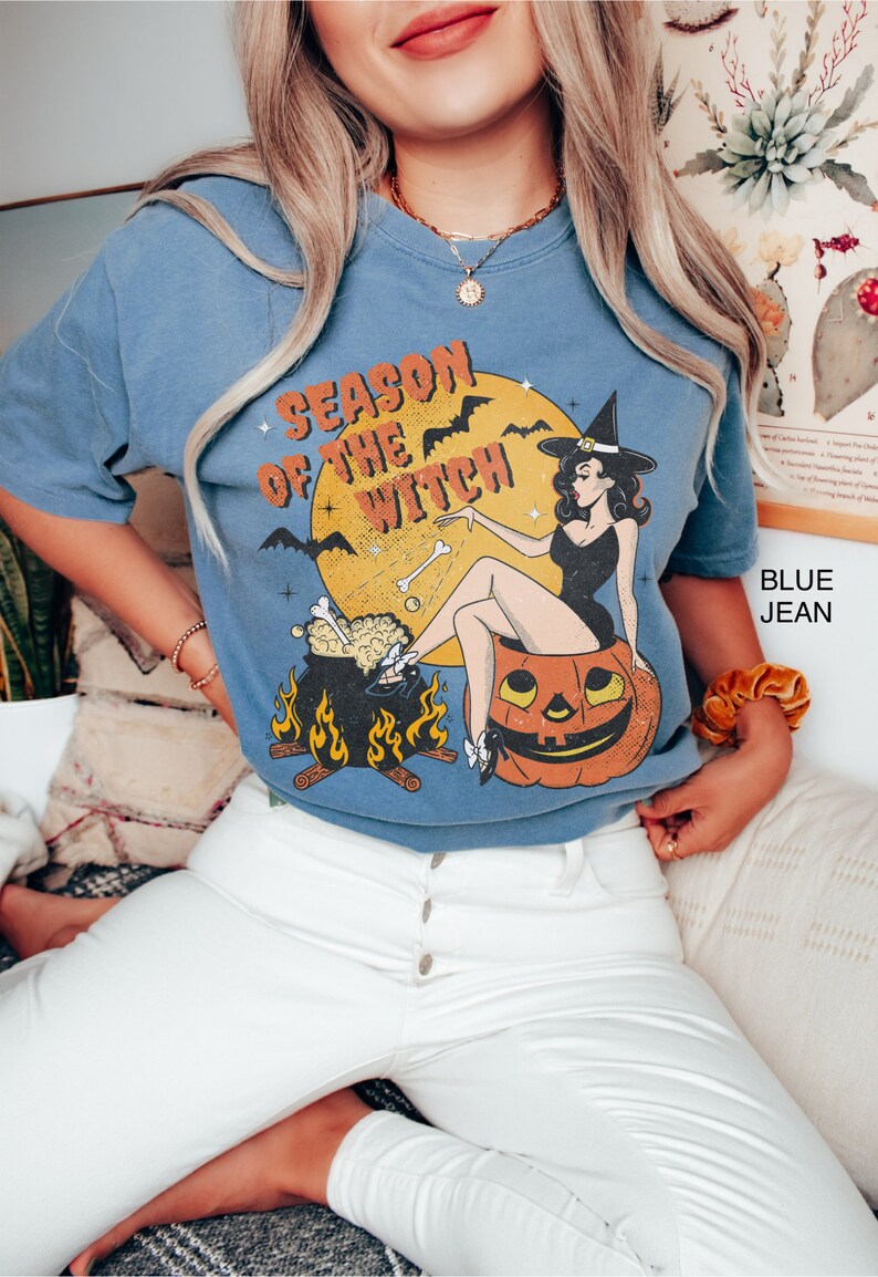 Puede incluir: Una camiseta azul con una ilustraci&oacute;n vintage de una bruja sentada sobre una calabaza. El texto "Season of the Witch" est&aacute; impreso en la camiseta.
