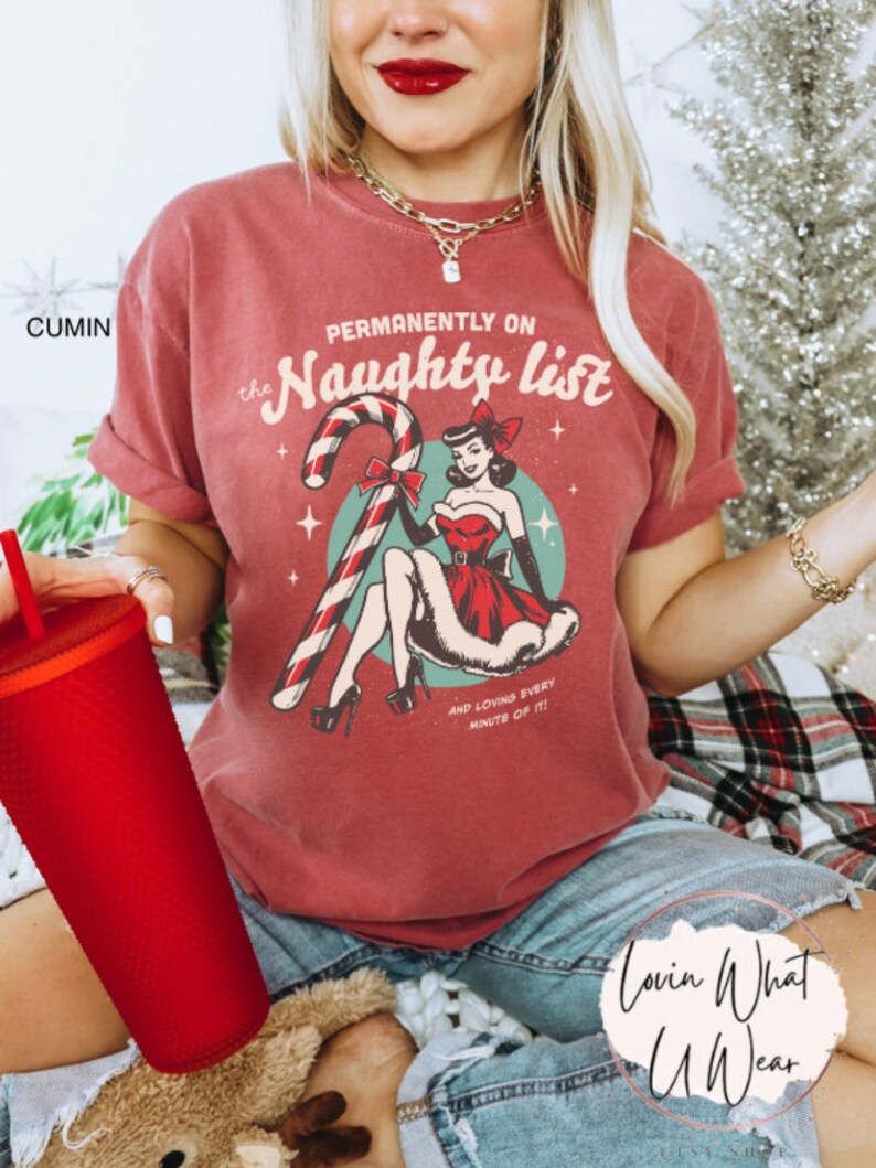 May include: A red t-shirt with a vintage-style illustration of a woman in a red dress and a candy cane. The text "Permanently on the Naughty List" is printed on the shirt. The text "And loving every minute of it!" is printed below the illustration.