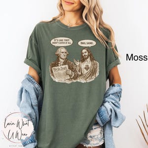 Może przedstawiać: Zielona koszulka w kolorze mchu z grafiką George'a Washingtona i Jezusa z dymkami. Washington trzyma dokument z napisem "We the People". Dymki m&oacute;wią "It's like they didn't listen at all" i "Bro, same."