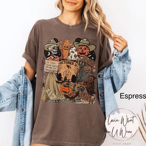 May include: Espresso-colored t-shirt with a Halloween-themed graphic. The design features a skeleton, a pumpkin, and other spooky characters. Text includes "October 31" and "Halloween Never Die". The shirt is part of a clothing collection.