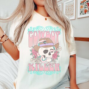 Op de afbeelding: Een lichtgeel T-shirt met een grafische print van een schedel die een cowboyhoed en een slang draagt. De tekst "COWBOY KILLER" is gedrukt in roze en blauwe letters.