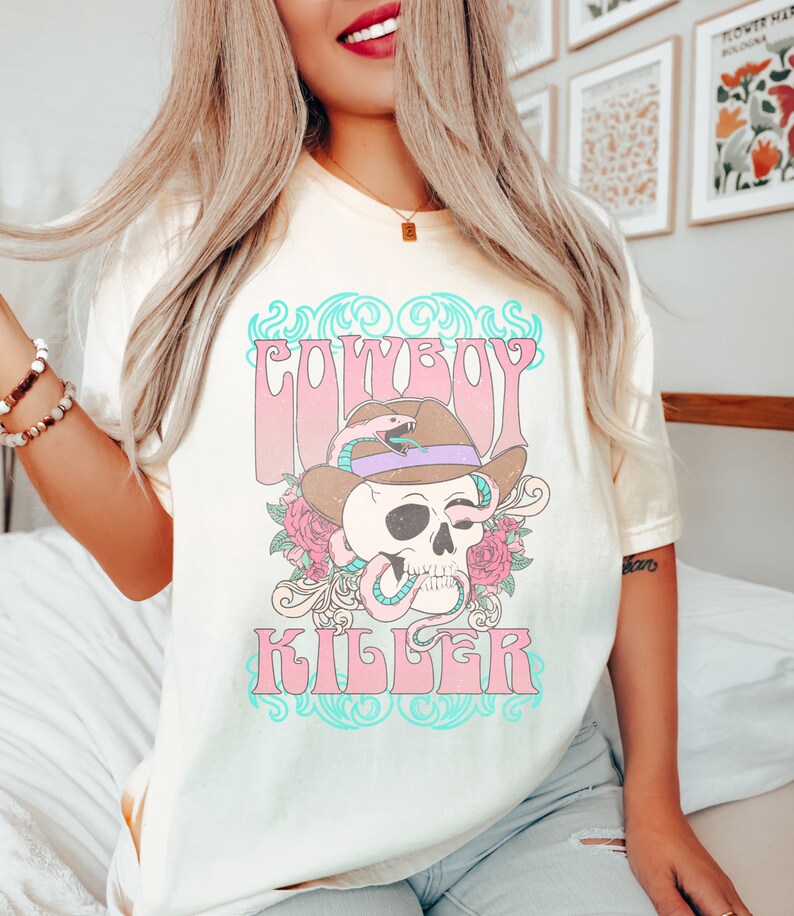 Op de afbeelding: Een lichtgeel T-shirt met een grafische print van een schedel die een cowboyhoed en een slang draagt. De tekst "COWBOY KILLER" is gedrukt in roze en blauwe letters.