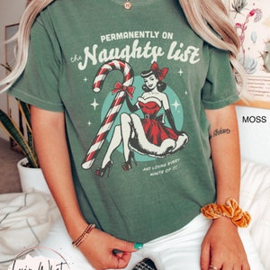 May include: A green t-shirt with a vintage-style graphic of a woman holding a candy cane. The text on the shirt reads "Permanently on the Naughty List" and "And loving every minute of it!"