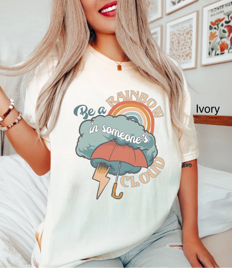 May include: A white t-shirt with a graphic design of a rainbow, a cloud, an umbrella, and lightning. The text on the shirt reads "Be a rainbow in someone's cloud".