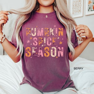 Może przedstawiać: Koszulka w kolorze malwy z napisem "Pumpkin Spice Season" w kolorze pomarańczowym, r&oacute;żowym i białym z gwiazdami wok&oacute;ł napisu.