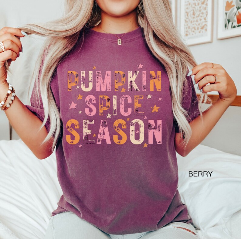 Może przedstawiać: Koszulka w kolorze malwy z napisem "Pumpkin Spice Season" w kolorze pomarańczowym, r&oacute;żowym i białym z gwiazdami wok&oacute;ł napisu.