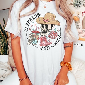 Pu&ograve; includere: Maglietta bianca con una grafica di uno scheletro che indossa un cappello da cowboy e tiene una tazza di caff&egrave; freddo. Il testo "Caffeine and Chaos" &egrave; scritto intorno allo scheletro.