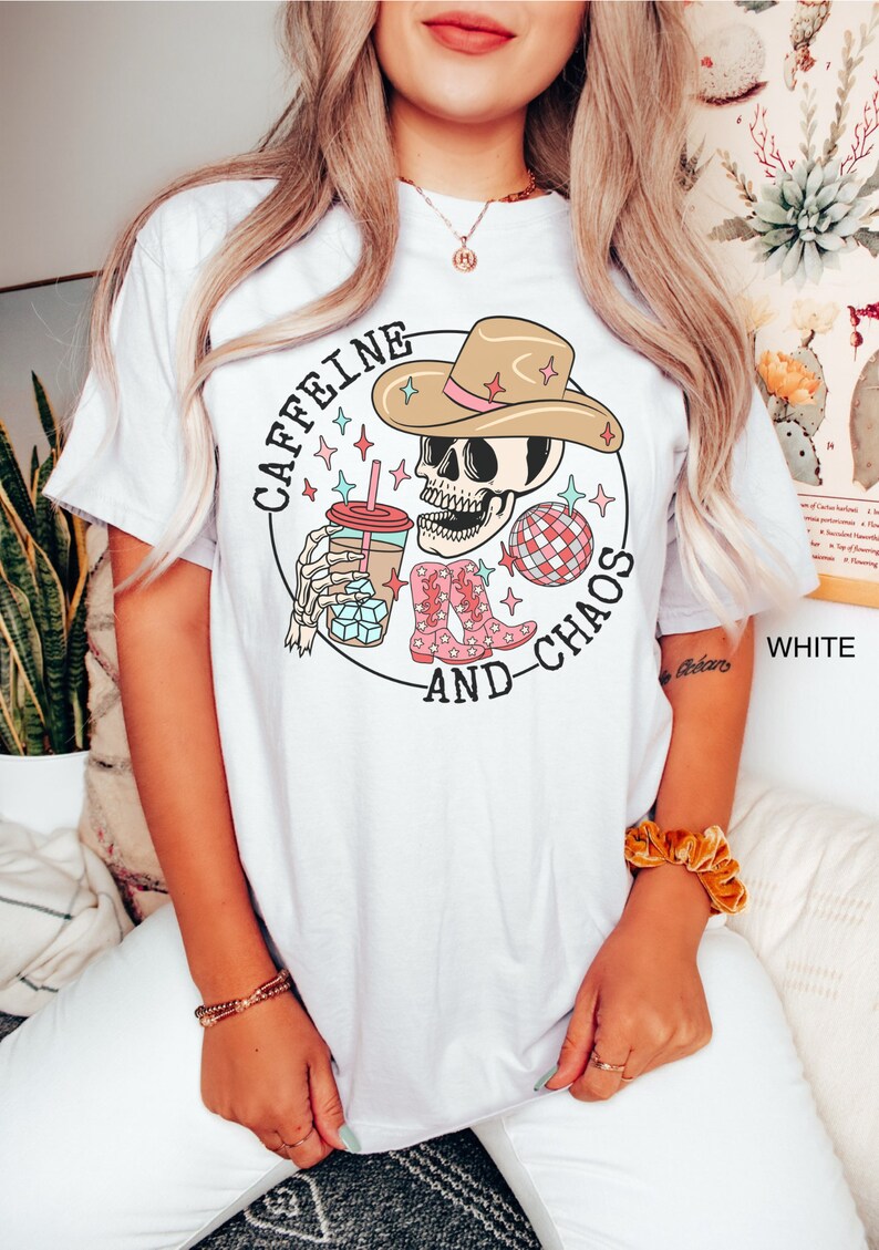 Pu&ograve; includere: Maglietta bianca con una grafica di uno scheletro che indossa un cappello da cowboy e tiene una tazza di caff&egrave; freddo. Il testo "Caffeine and Chaos" &egrave; scritto intorno allo scheletro.